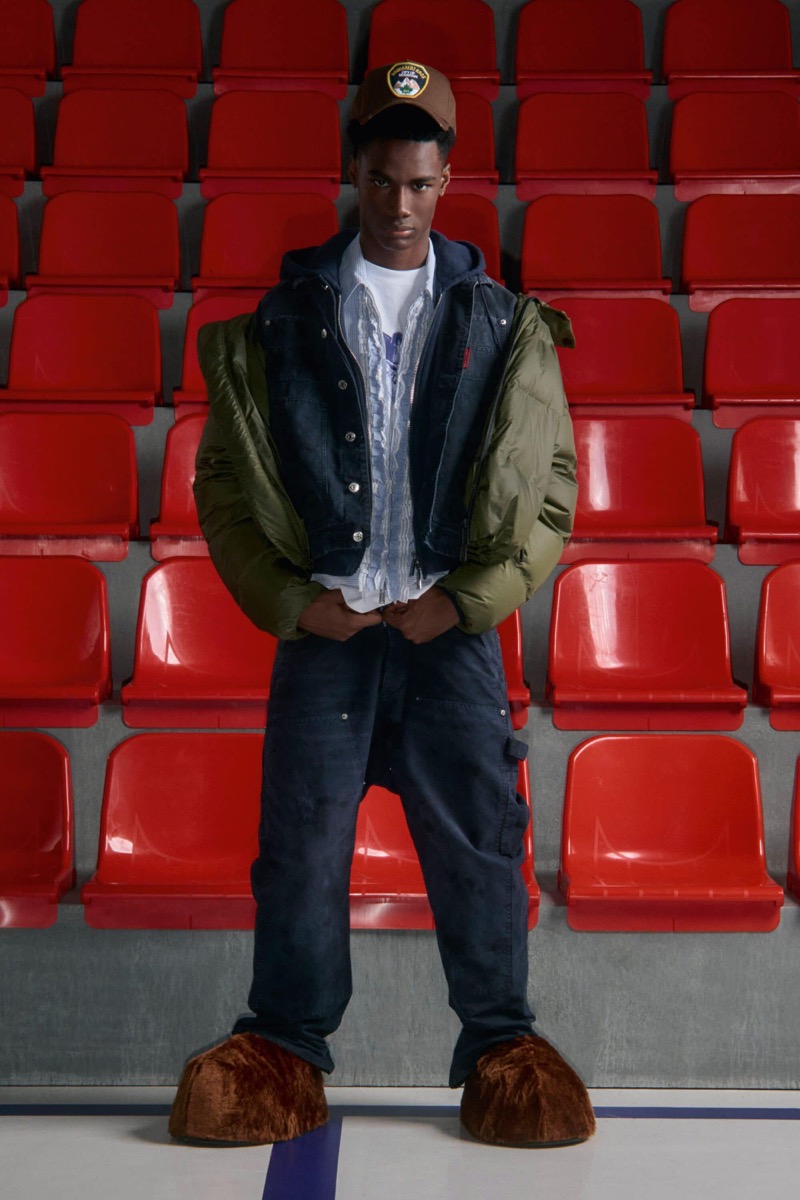 Nehemiah Kargbo wears a brown patch baseball cap and a denim trucker vest over an olive green down puffer and white tee with navy carpenter pants and brown faux fur clog mules