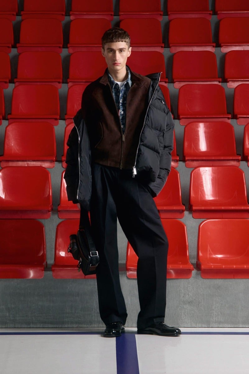 Madson Santos wears a brown corduroy zip jacket with a black down puffer draped over the shoulders, a printed shirt, black wool trousers, and black derby shoes, holding a small black leather bag