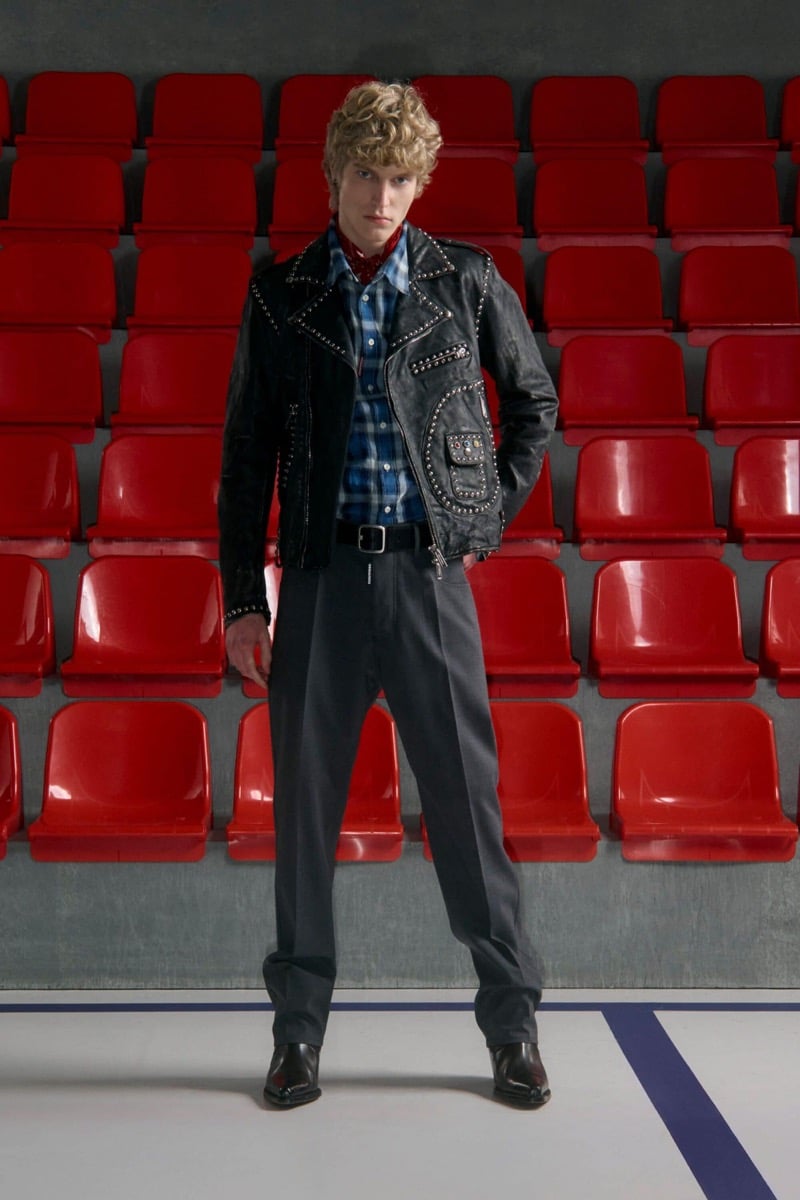 Kristaps Kallings wears a studded black leather biker jacket over a blue plaid flannel shirt with a red bandana, grey wool trousers, and black Western boots