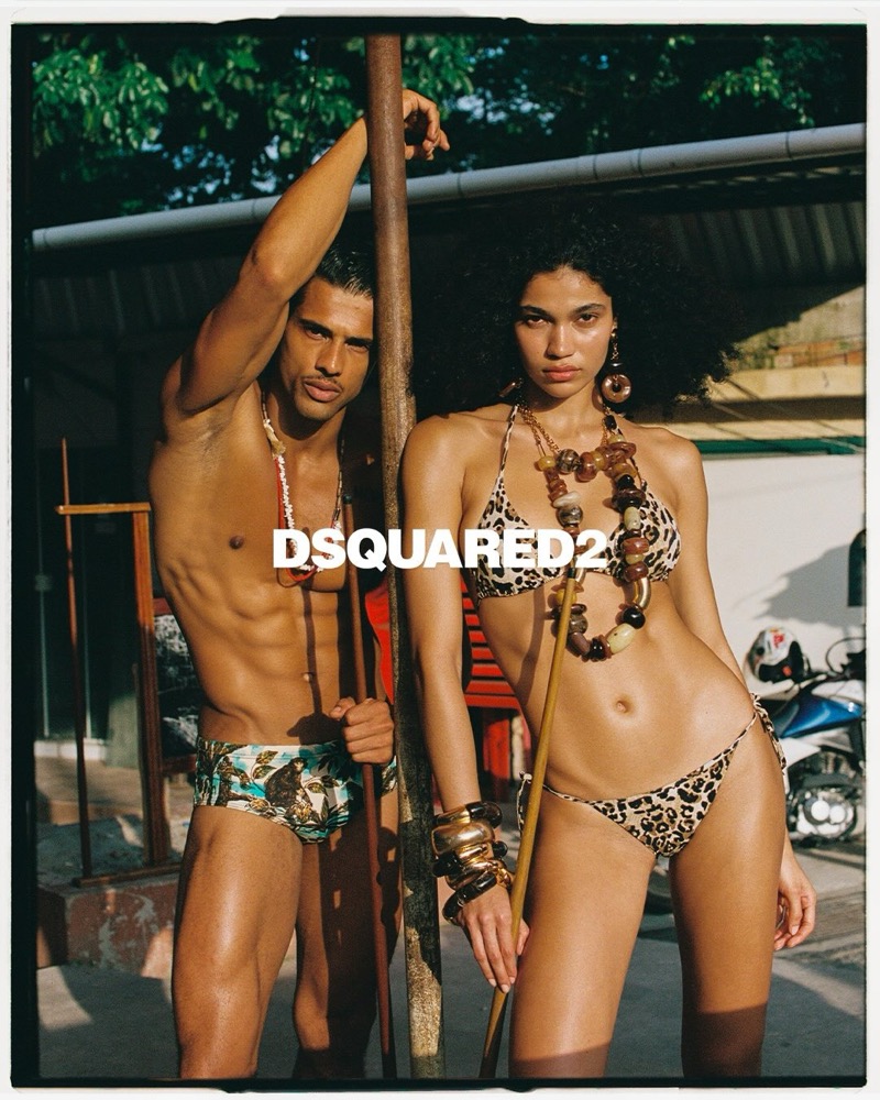Jonas in printed Dsquared2 swim briefs leans on a wooden pole beside a woman in a leopard-print Dsquared2 bikini and stacked amber-bead necklaces, the Dsquared2 logo set across the frame