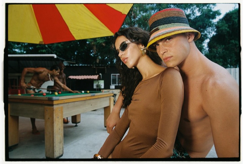 A woman in narrow sunglasses and a fitted brown long-sleeve top sits back-to-back with a shirtless man in a striped crochet bucket hat beneath a red-and-yellow striped umbrella, an outdoor pool table behind them