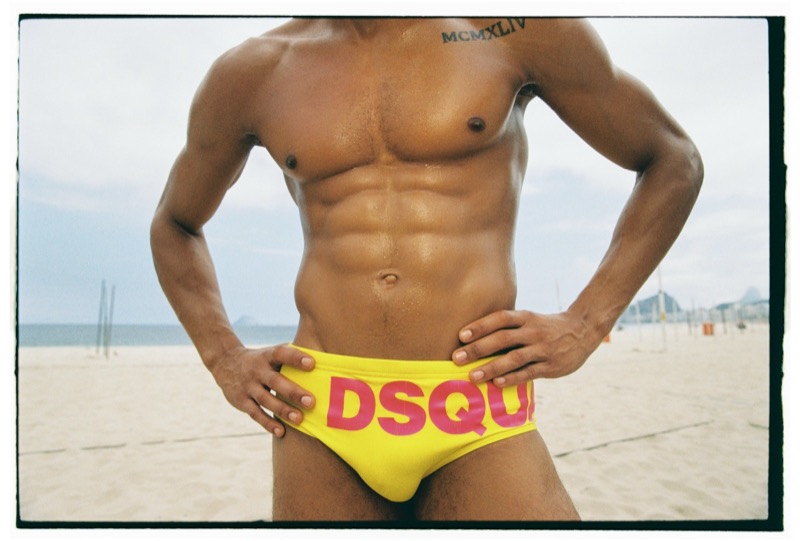 Cropped torso shot of Jonas on Copacabana beach in yellow Dsquared2 logo swim briefs, hands resting at his waist, his MCMXLIV chest tattoo visible