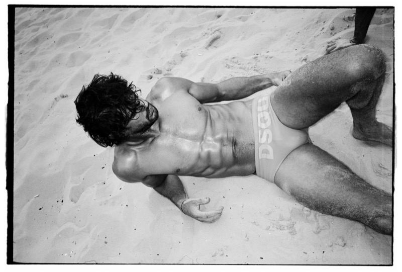Overhead black-and-white shot of Marlon lying back in the sand in white Dsquared2 logo swim briefs