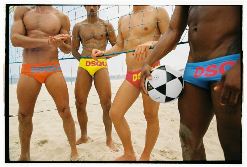 Four shirtless men stand at a beach volleyball net in coordinating Dsquared2 logo swim briefs — orange, yellow, red, and blue —