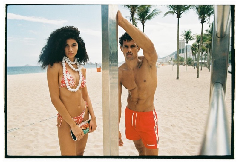 A woman in a red-and-white printed Dsquared2 bikini and oversized white shell necklaces stands beside Jonas in red Dsquared2 swim shorts at a stainless-steel Copacabana beach shower