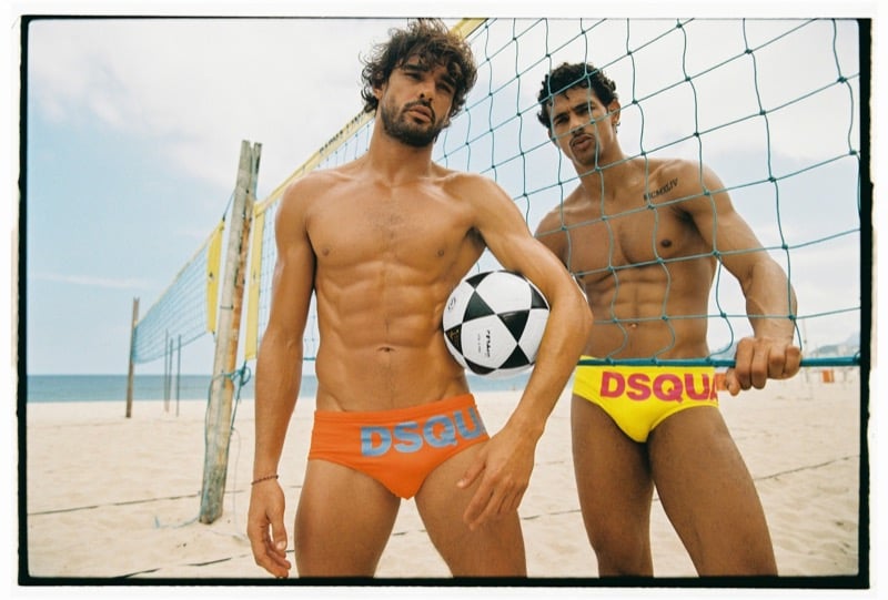 Marlon and Jonas stand at a beach volleyball net holding a soccer ball, Marlon in orange Dsquared2 swim briefs with a gray logo and Jonas in yellow briefs with a pink logo