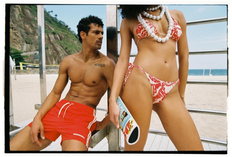 Jonas in red Dsquared2 swim shorts sits on a beach railing as a woman in a red Coca-Cola-print Dsquared2 bikini and chunky white bead necklace stands in the foreground holding a can