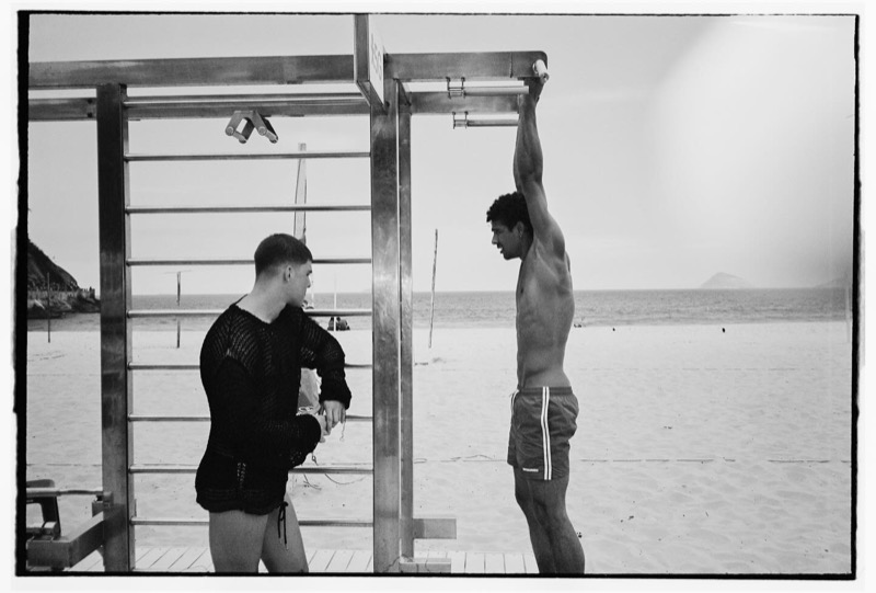 Two men at a beach pull-up rig on Copacabana, one in a black knit mesh top and one in red Dsquared2 swim shorts stretching upward, photographed in black and white