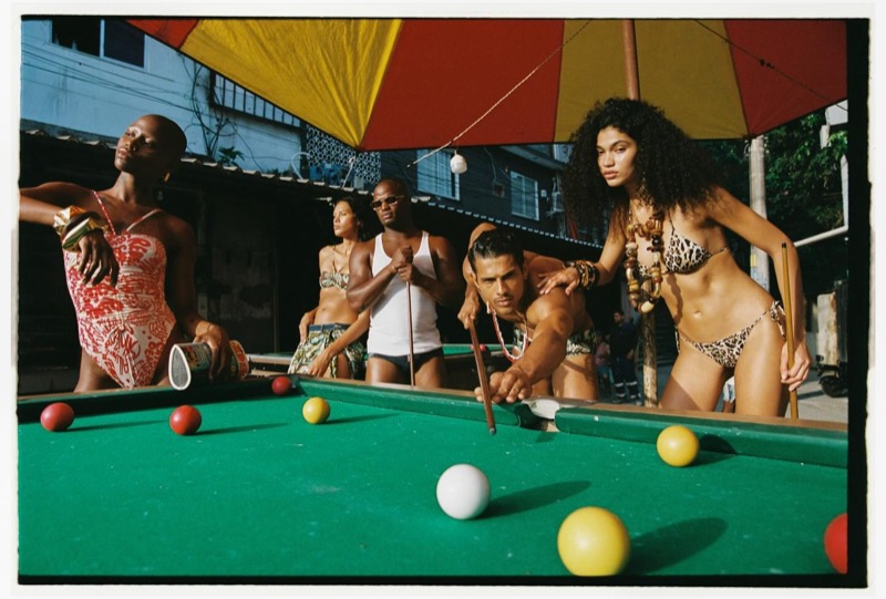 A group of models gathered around an outdoor pool table beneath a red-and-yellow striped umbrella