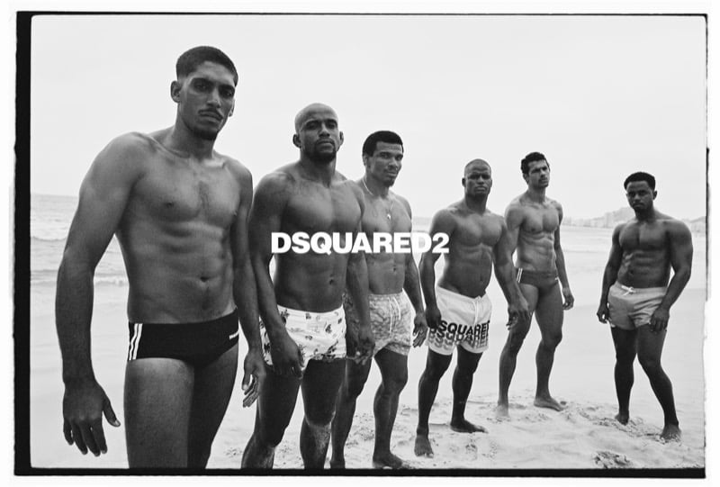 A line of six shirtless men in Dsquared2 swim shorts and briefs stand along a Rio beach