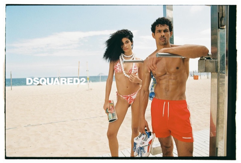 A woman in a red printed Dsquared2 bikini and white shell necklaces holds a glass bottle beside Jonas in red Dsquared2 swim shorts leaning on a beach shower rail, the Dsquared2 logo set across the sky