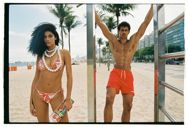 A woman in a red-and-white printed Dsquared2 bikini and chunky white bead necklace stands beside Jonas in red Dsquared2 shorts who hangs from a stainless-steel pull-up rig on Copacabana