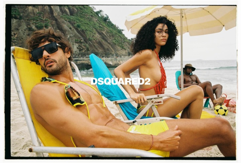 Marlon in dark sunglasses reclines on a yellow beach chair in yellow Dsquared2 swim shorts beside a woman in a red crocheted bikini top