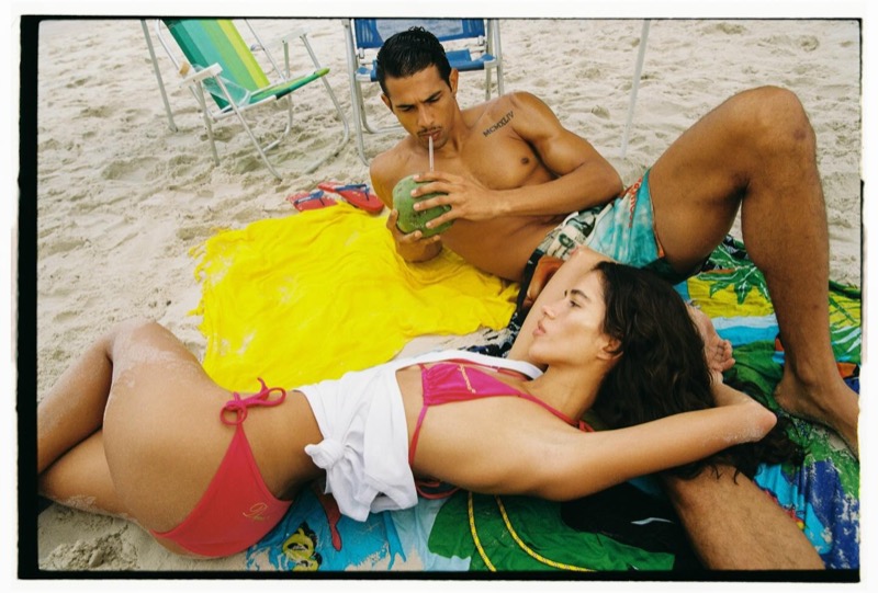 Jonas drinks from a green coconut while a woman in a pink Dsquared2 bikini and white tee reclines beside him