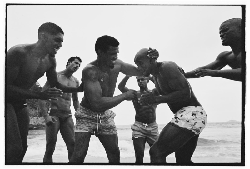 A group of shirtless men in Dsquared2 swim shorts and briefs laugh and grapple at the water’s edge along a Rio beach, photographed in black and white