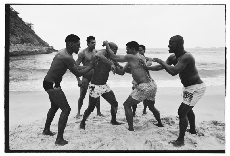 A group of shirtless men in Dsquared2 swim shorts and briefs grapple playfully in a circle on a Rio beach