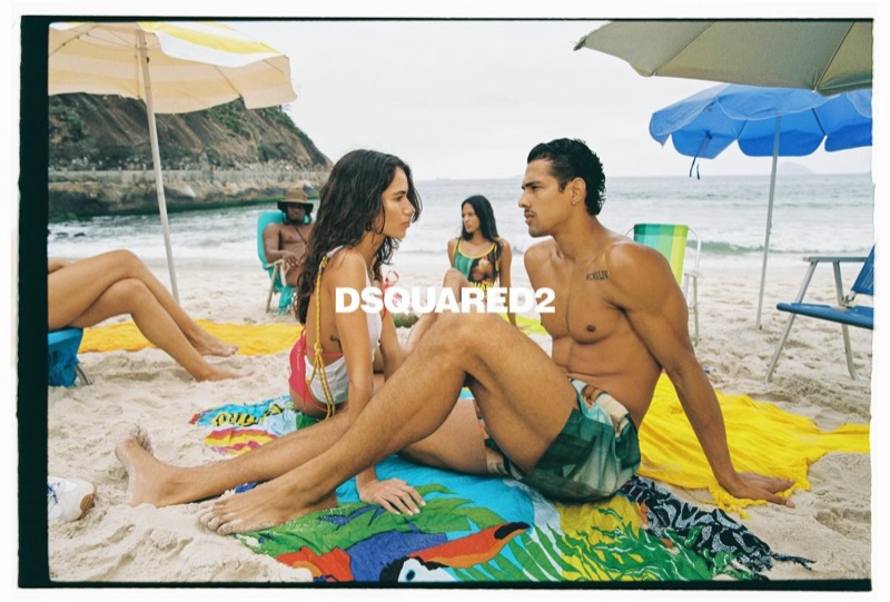 Jonas in printed Dsquared2 swim shorts sits across a colorful printed beach towel from a woman in a white tank and printed bikini bottoms