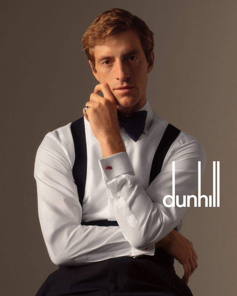 Orfeo poses in a white tuxedo shirt with grosgrain braces, a bow tie, and pleated trousers, resting his chin on his hand with a silver pinky ring