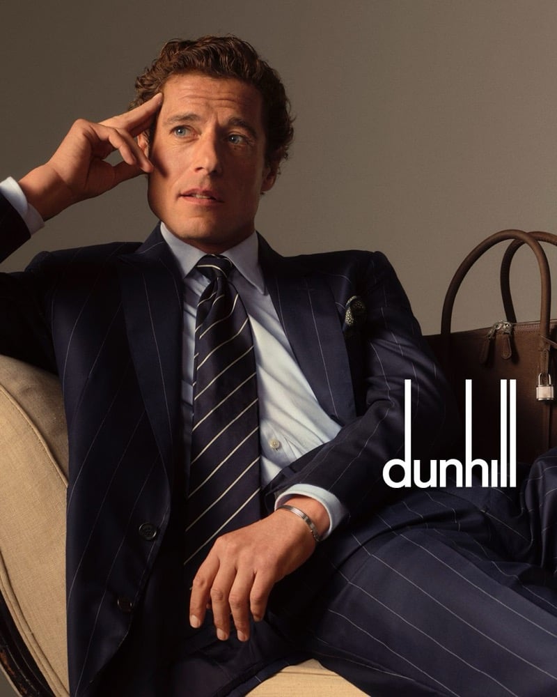 Jon Paul reclines in a navy pinstripe suit with a light blue shirt, a navy and silver striped tie, and a silver bracelet, beside a brown leather Dunhill holdall