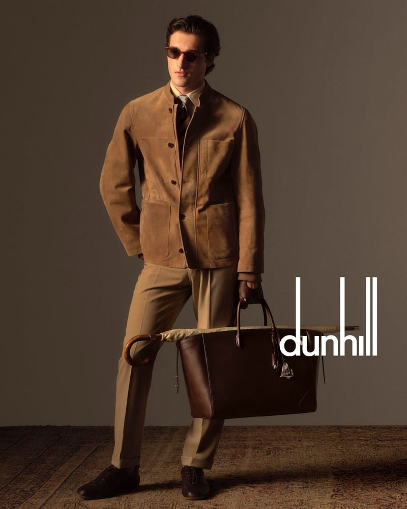 Adam wears tortoise sunglasses, a tan suede shirt jacket over a white shirt with a dark tie, and trousers, carrying a large brown leather tote and a wood-handled umbrella