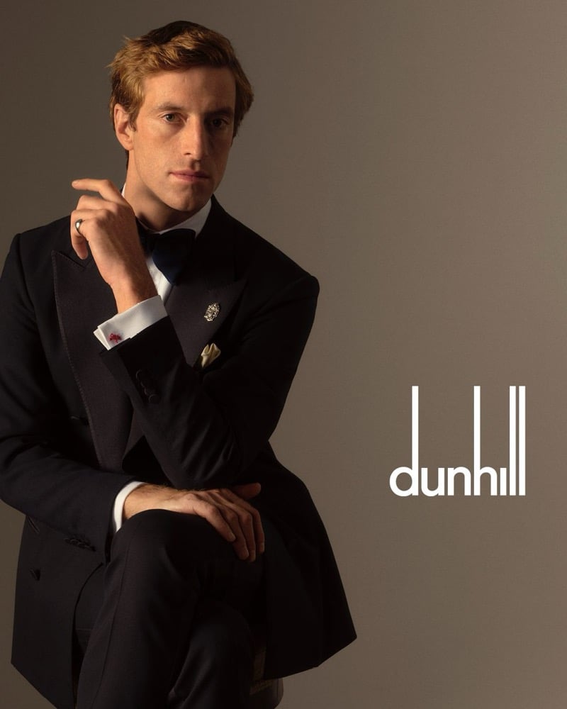 Orfeo sits in a black peak-lapel dinner jacket with grosgrain trim, a white shirt, a bow tie, a lapel crest pin, and a cream pocket square