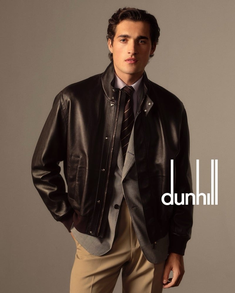 Adam wears a dark brown lambskin bomber jacket over a gray blazer, a lilac shirt, a brown striped tie, and tan trousers