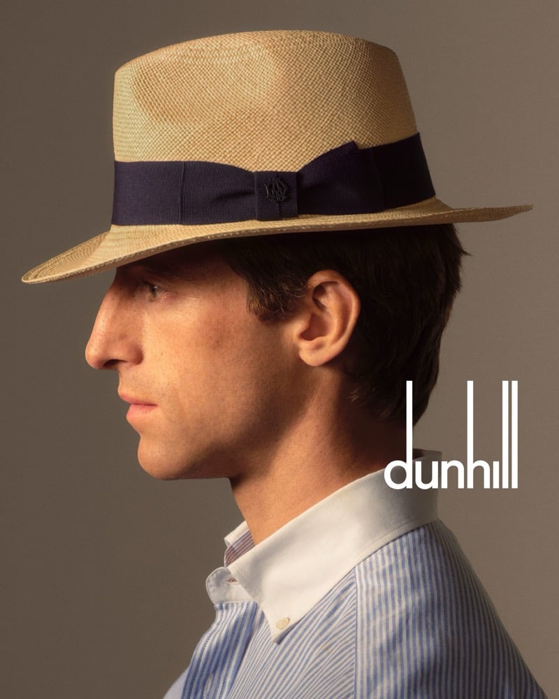 Orfeo in profile wearing a natural straw Panama hat with a navy grosgrain band and crest detail, over a blue stripe shirt with a white contrast collar