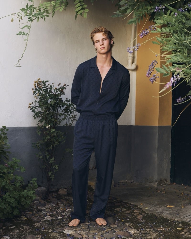 Henry Rank stands barefoot on stones in a Mediterranean courtyard surrounded by wisteria and greenery, a chalk-white facade above a dark gray lower wall behind him, wearing a navy jacquard-pattern Emporio Armani camp-collar long-sleeve shirt left half-open over matching navy drawstring pajama trousers, blonde tousled hair