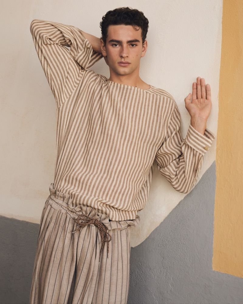 Jack Wardill leans against a color-blocked Mediterranean wall in chalk-white and pale gray with a yellow column at the edge, one hand raised flat to the surface, wearing a cream and tan vertical-stripe Emporio Armani boatneck top with wide-cut beige and brown striped drawstring linen trousers, dark curly hair