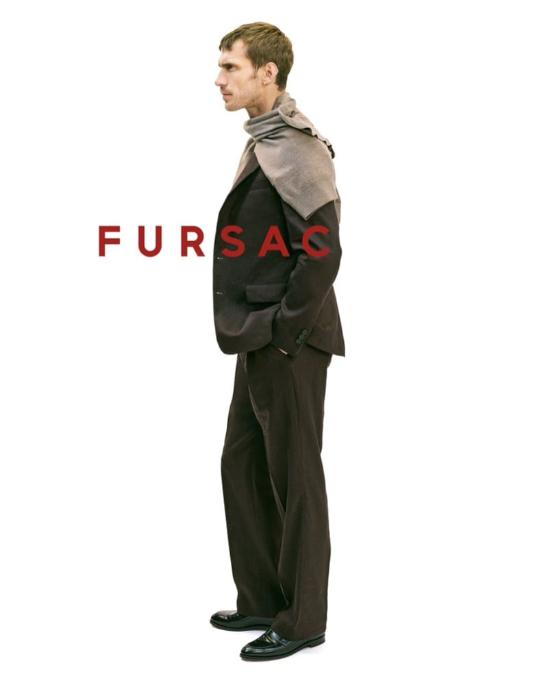Side profile of Clément in a dark brown suit with a taupe knit on his shoulders, the trousers breaking long over polished black loafers