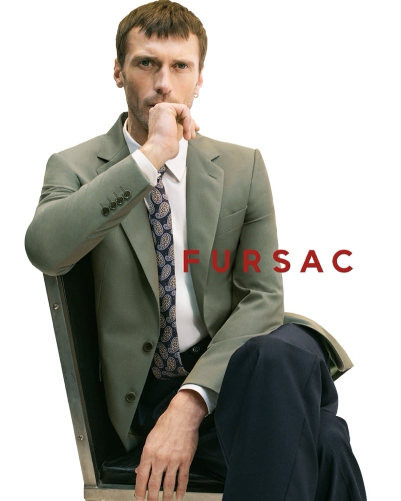 Clément sits in a wire chair in a sage green blazer over a white shirt with a navy paisley tie and dark navy trousers