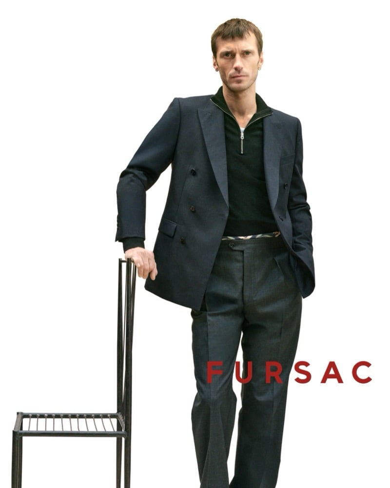 Clément leans on a wire chair in a navy double-breasted suit worn over a black quarter-zip knit zipped high against the neck, with charcoal trousers