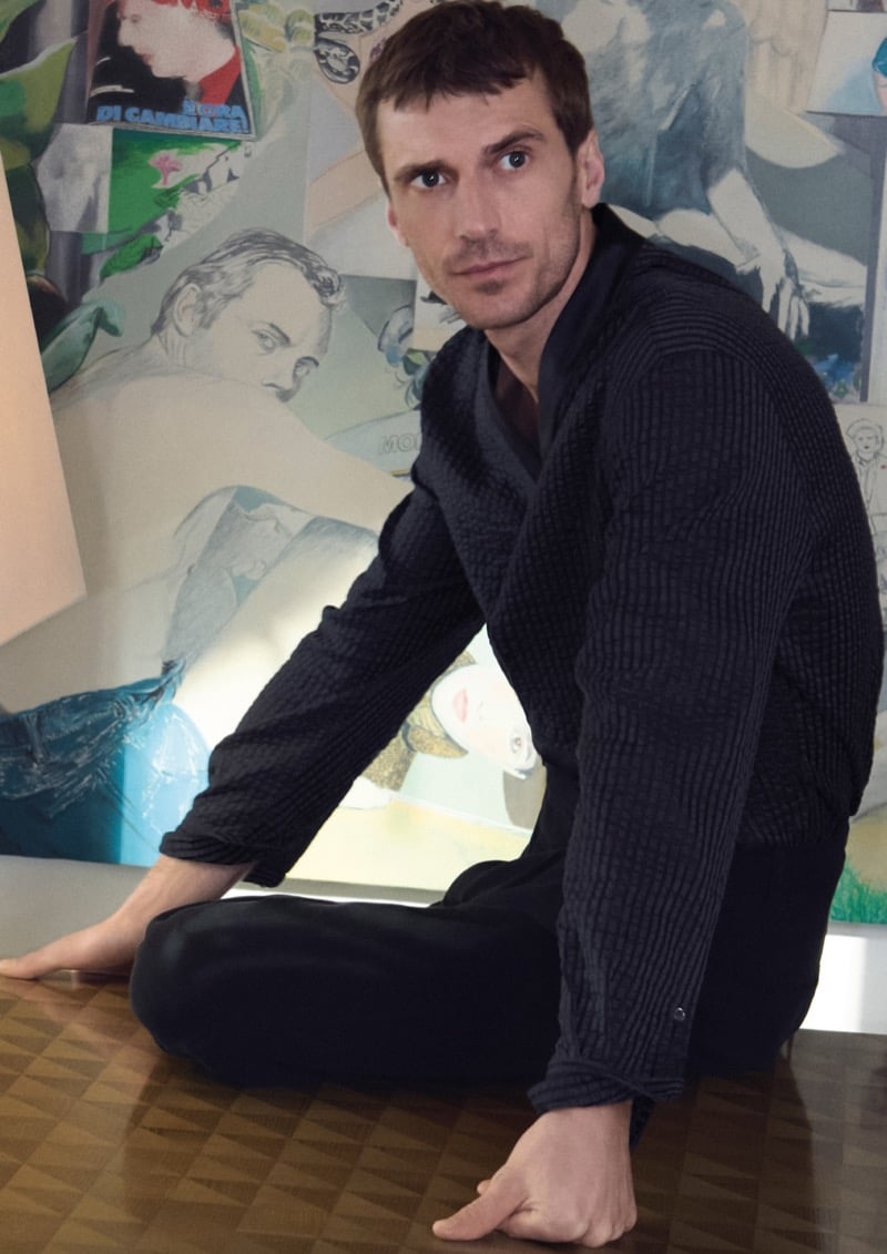 Clément sits on a parquet table in a deconstructed dark gray textured zip jacket and dark trousers in front of a layered painting