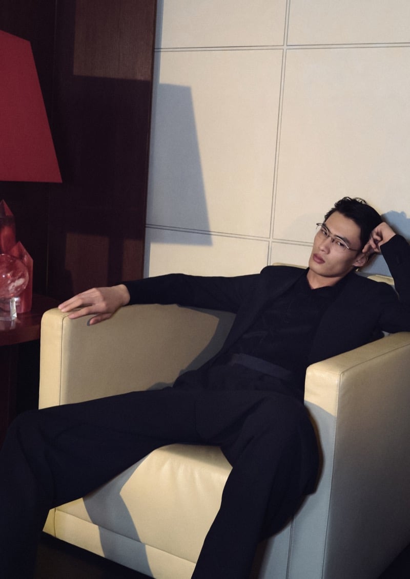 Zhaoyi reclines in a cream leather armchair beside a red lamp in a soft-shouldered black Giorgio Armani suit and clear-frame glasses