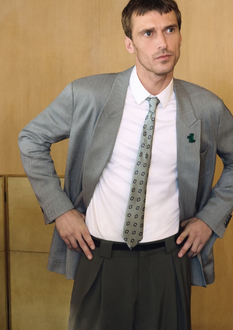 Clément stands with hands on hips in a relaxed light gray Giorgio Armani blazer over a white shirt with a patterned tie tucked into dark gray pleated trousers