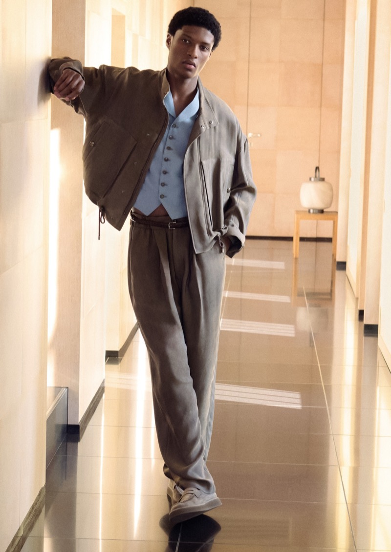 Aboubakar leans against a sunlit hallway wall in a taupe Giorgio Armani bomber jacket over a pale blue waistcoat with wide-leg taupe trousers and suede shoes