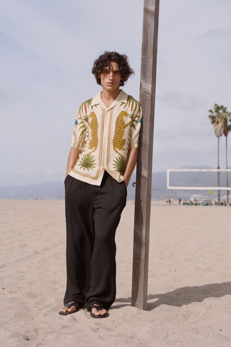 Morgan stands against a wooden volleyball pole on Venice Beach in a cream camp collar shirt patterned with rampant cheetahs, palms, and bordered stripe work, plain black linen pants, and brown leather thong sandals