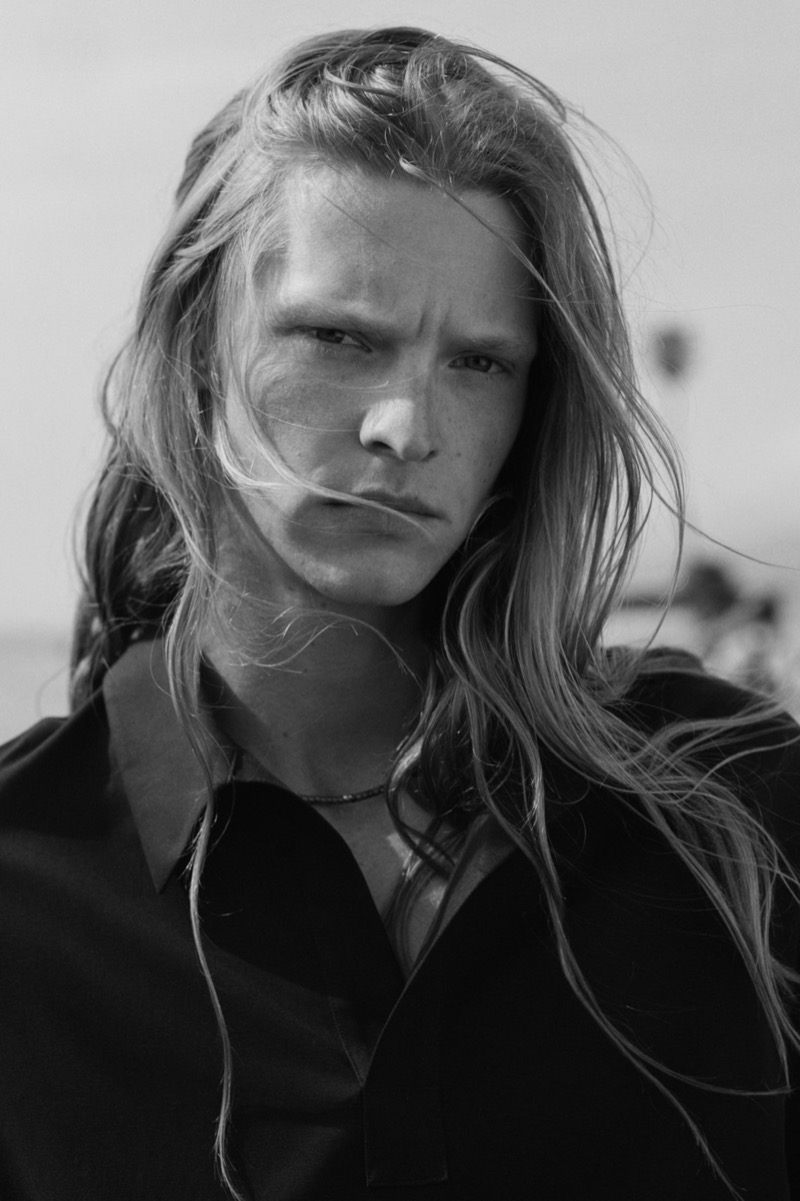 Black and white close-up portrait of Luke with long blonde hair blowing across his face on Venice Beach, the collar of a chocolate popover shirt visible at his shoulders