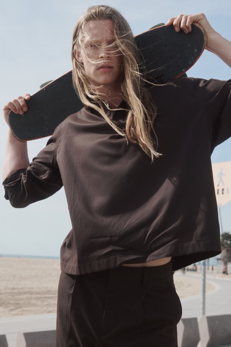 Luke stands on the Venice boardwalk holding a skateboard balanced across his shoulders in a chocolate brown popover shirt with dark slouched trousers, the sand and bay behind him