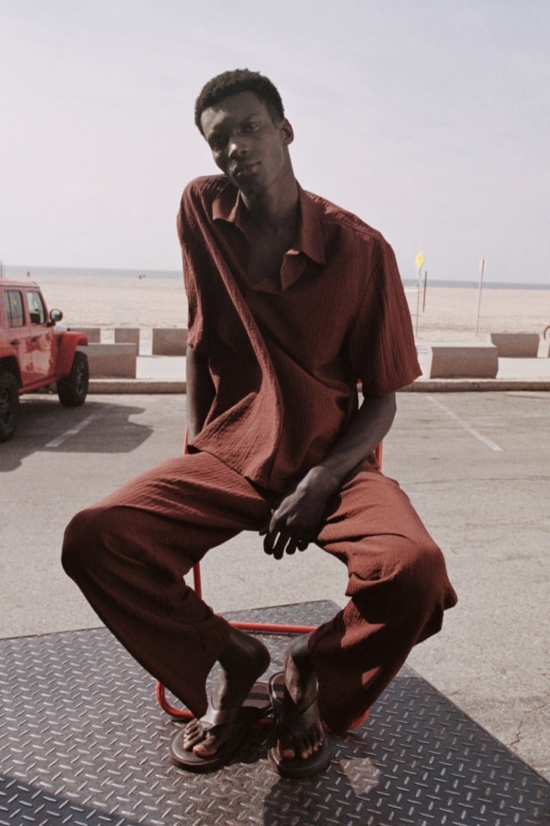 Momo sits on a red chair on a diamond-plate platform near the Venice boardwalk in a matching terracotta crinkled cotton camp collar shirt and trousers with dark leather thong sandals, a red Jeep and the sand visible behind him
