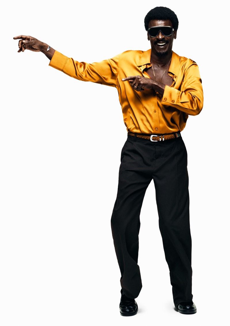 Seu points and laughs in a marigold satin shirt, braided belt, and black pleated trousers with sport sunglasses against a white backdrop for H&M