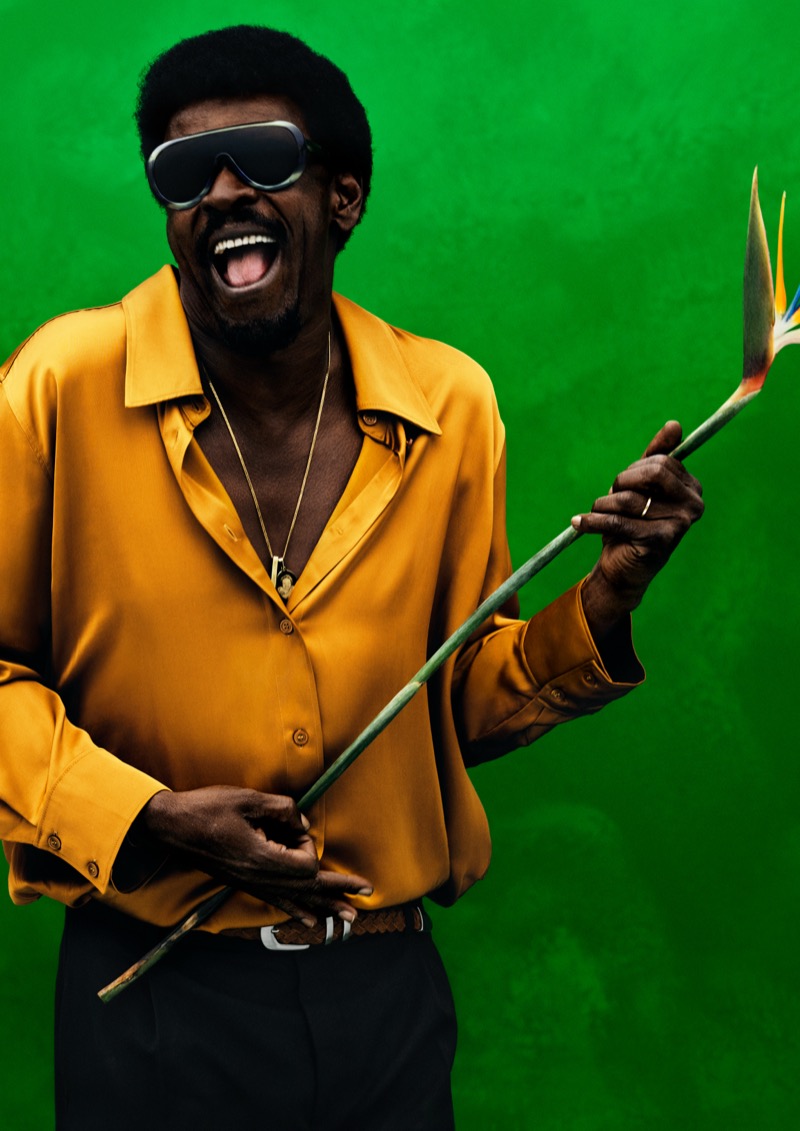 Seu laughs against a green backdrop in an open marigold satin shirt and goggle sunglasses, holding a bird-of-paradise flower