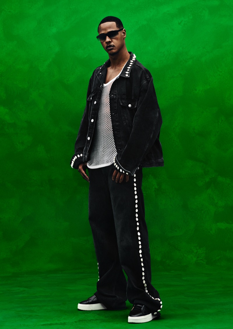 TZ stands against a green wall in a black denim jacket and matching wide-leg jeans trimmed with cowrie shells, layered over a white mesh tank with black sneakers