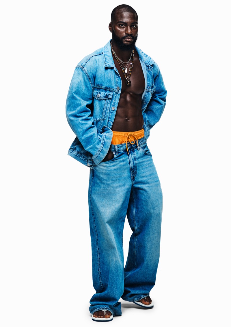Washington stands in an oversized blue denim jacket worn open over bare chest with layered beaded necklaces, wide-leg jeans, and orange swim shorts pulled above the waistband