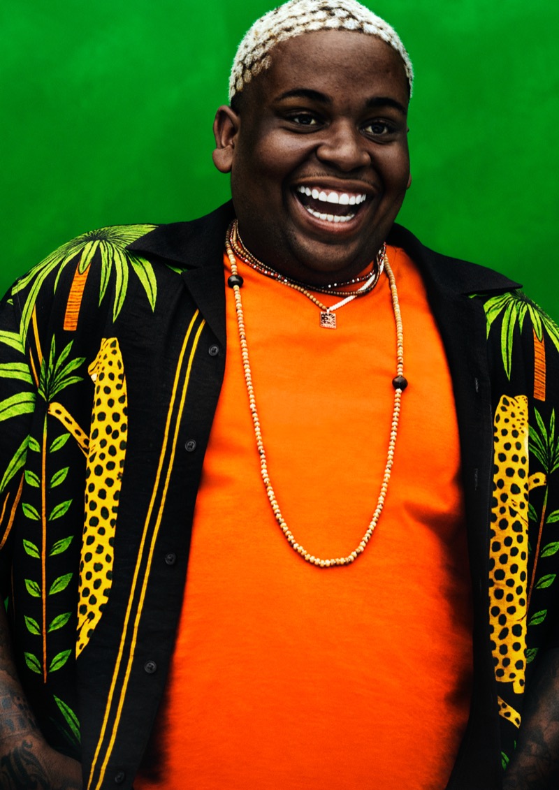 Close portrait of Ramon laughing against a green backdrop in a black short-sleeve shirt with green palms and yellow leopard print over an orange tee with layered beaded necklaces