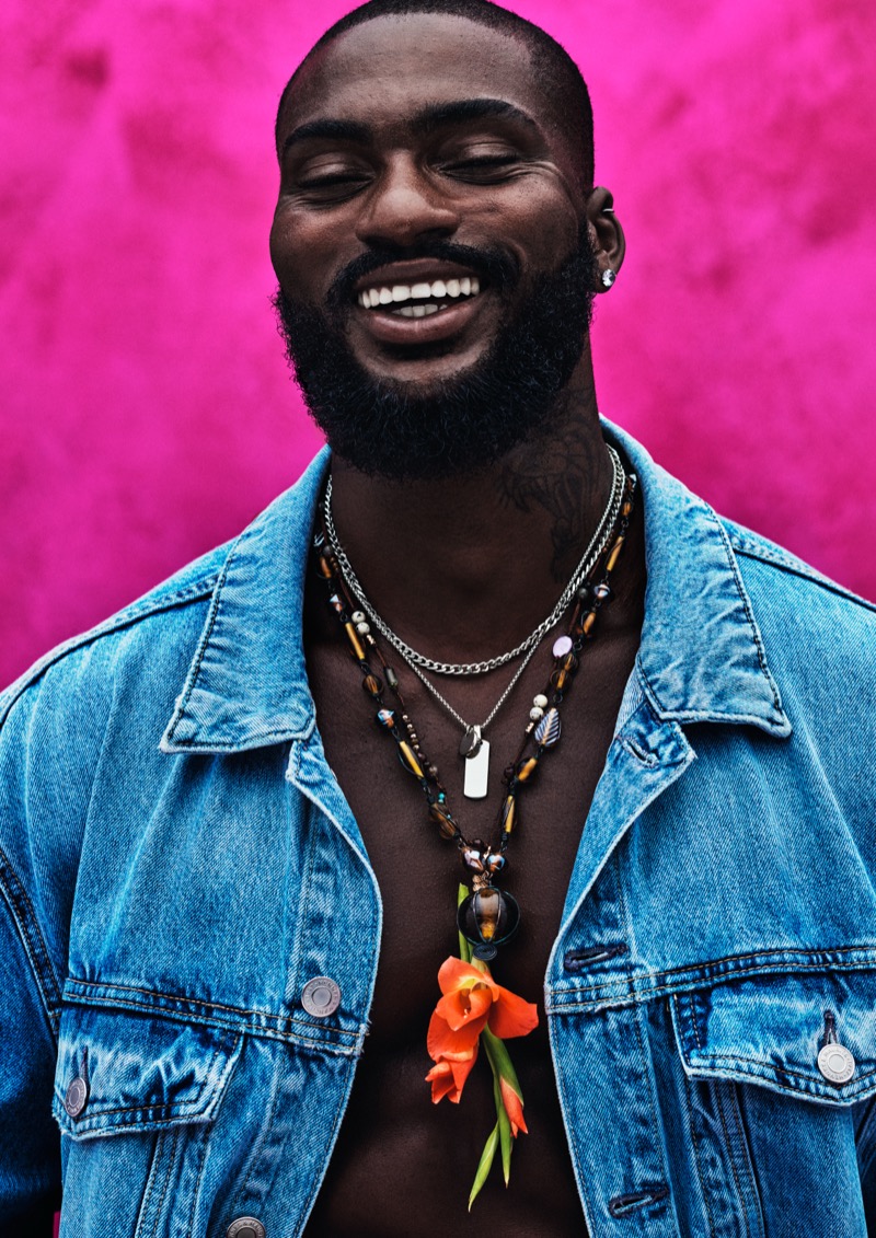 Close portrait of Washington smiling against a hot pink wall in an open blue denim jacket and stacked silver and beaded necklaces, with an orange gladiolus flower at the chest