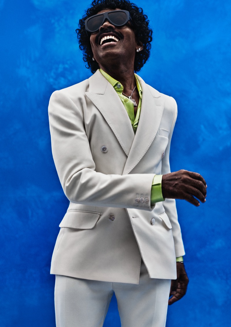 Indio laughs against a blue backdrop in an off-white double-breasted suit over a lime green satin shirt with sport sunglasses and a silver pendant necklace