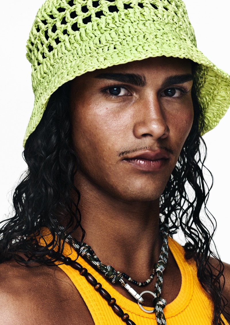 Close portrait of a young model with long wavy dark hair in a lime green crochet bucket hat, layered beaded and rope necklaces, and an orange ribbed tank
