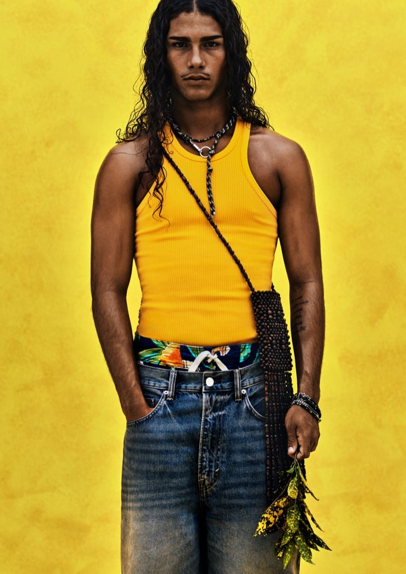 A model with long wavy dark hair stands against a yellow backdrop in an orange ribbed tank with layered beaded necklaces, a black beaded crossbody bag, and faded baggy jeans