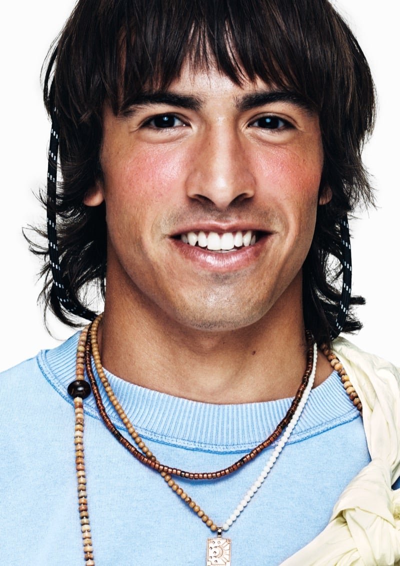 Close portrait of a smiling model with shaggy brown hair and bangs in a baby blue crewneck sweatshirt with stacked beaded necklaces and a printed scarf at the shoulder